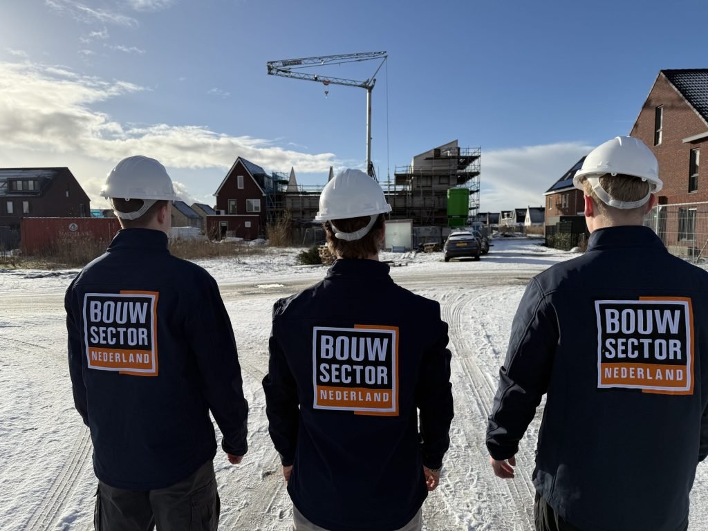 Drie vakmensen van Bouwsector Nederland met witte helm en bedrijfsjas op een nieuwbouwlocatie met een bouwkraan